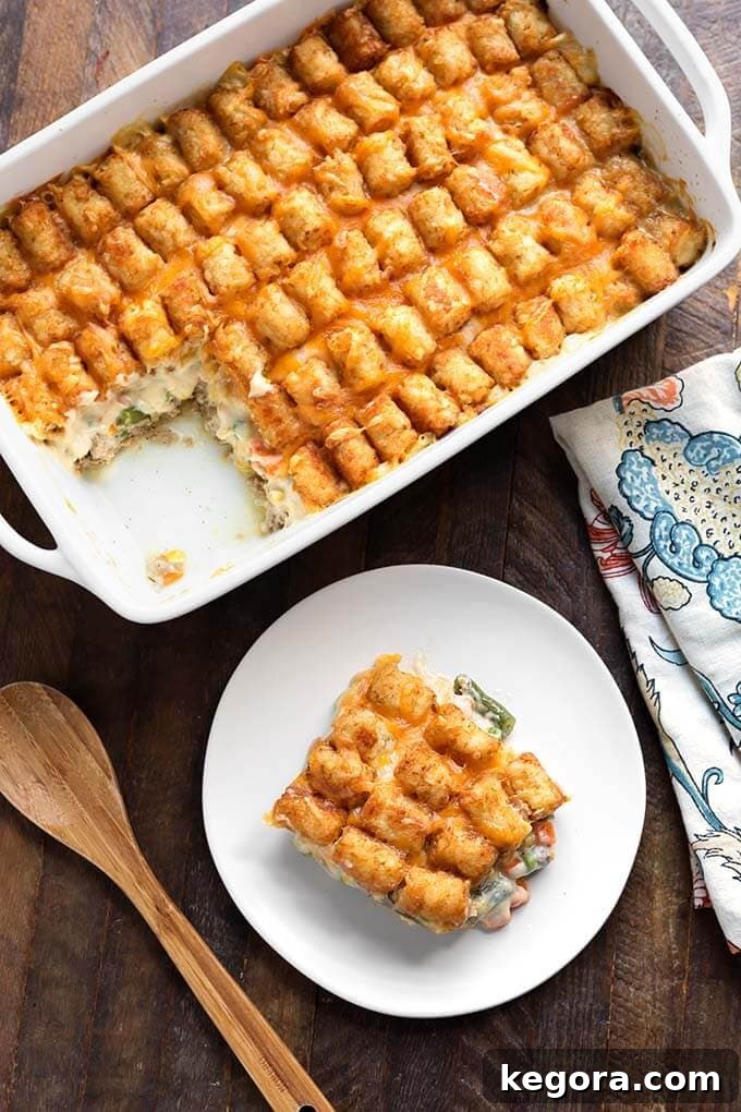 Slice of Hotdish Tater Tot Casserole on a white plate in front of remaining casserole in a baking dish