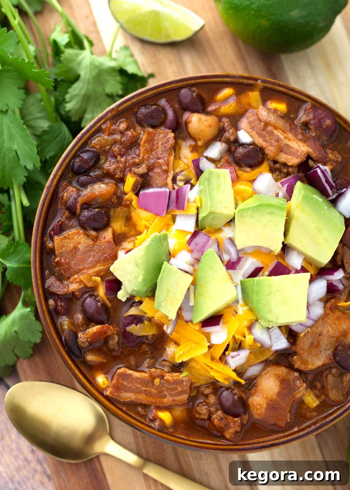 Beefy Bacon Chili in a brown bowl topped with cubed avocado, shredded cheese, and diced red onion