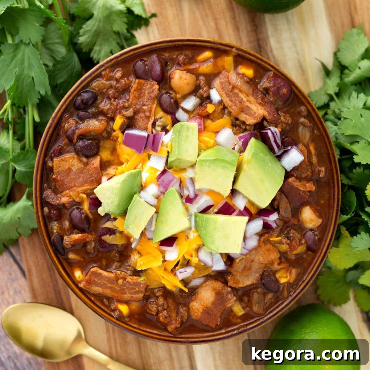 Beefy Bacon Chili in a brown bowl topped with cubed avocado, shredded cheese, and diced red onion