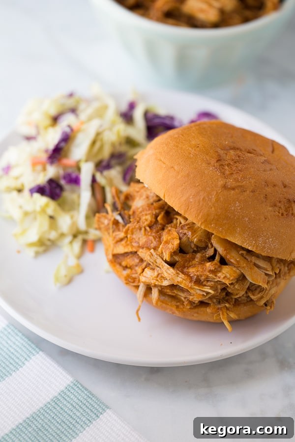 Shredded BBQ chicken served on a soft hamburger bun with a side of creamy coleslaw and a pickle