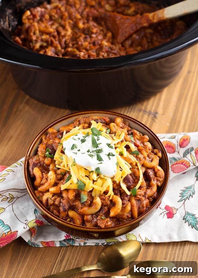 Chili Mac in a brown bowl topped with cheese and sour cream in front of a slow cooker