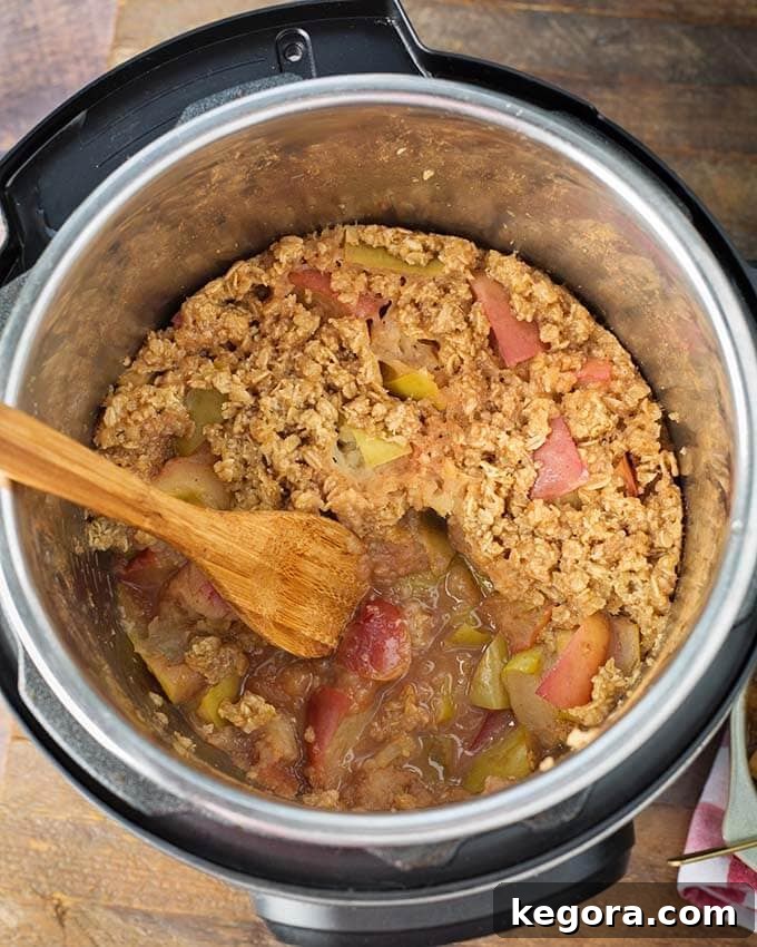 A pressure cooker pot filled with Instant Pot Apple Crisp, showcasing the cooked apples and topping, with a wooden mixing spoon resting inside.