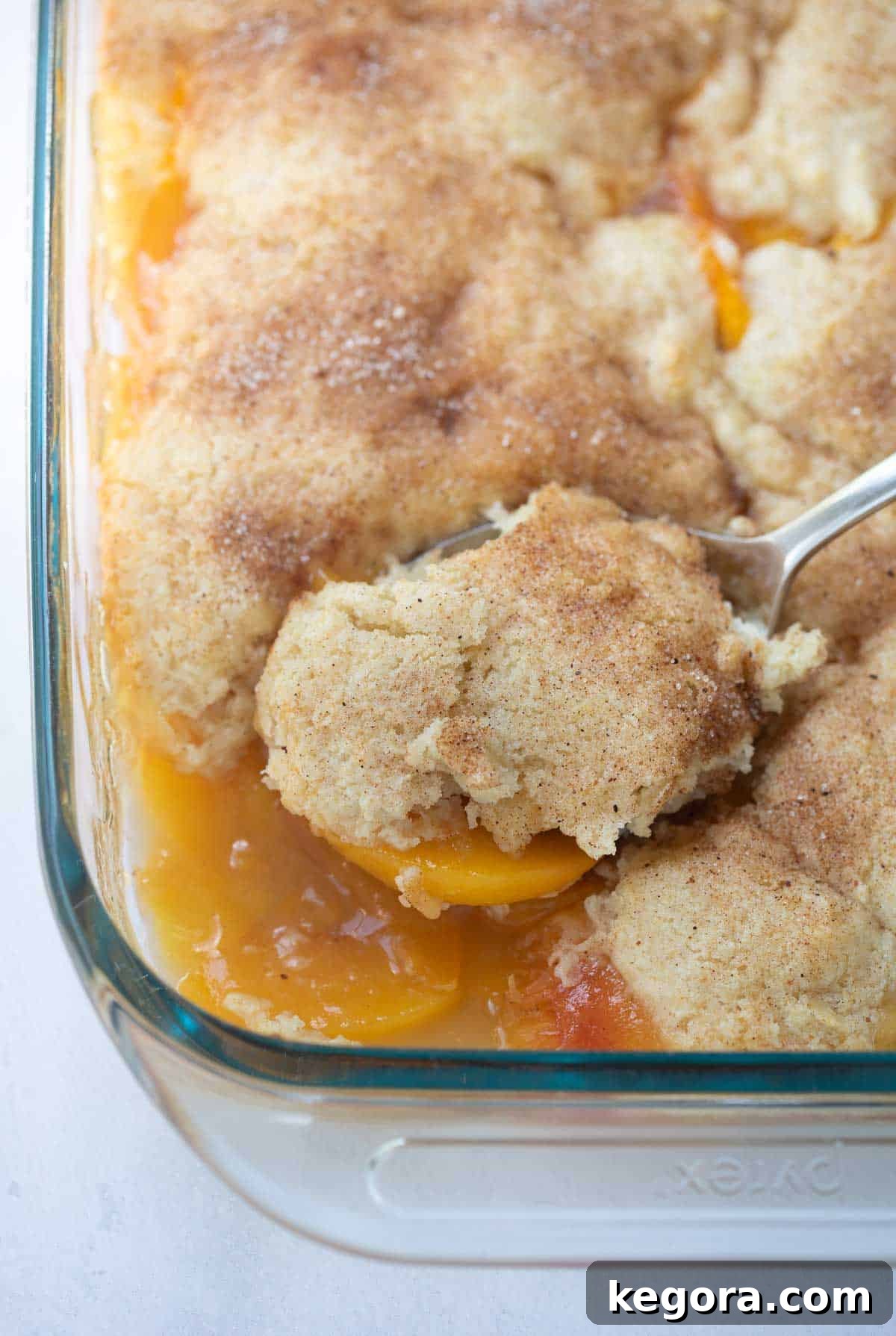 A scoop of homemade gluten-free peach cobbler being lifted from a baking pan, showing its juicy peach filling and golden-brown, fluffy topping.
