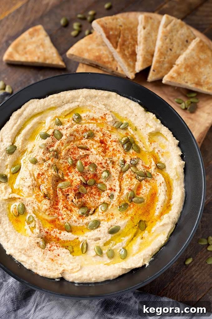 Hummus in a black bowl next to a wooden board with cut pieces of flatbread, garnished with paprika and pumpkin seeds