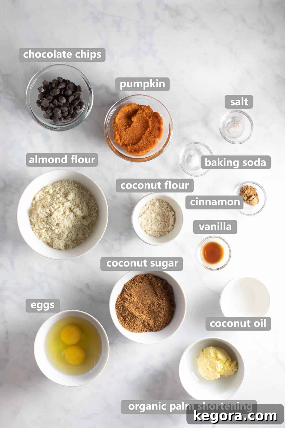 Bowls of ingredients including pumpkin puree, coconut sugar, palm shortening, and chocolate chips, arranged on a marble background for paleo pumpkin cookies recipe.