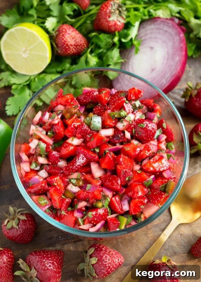 Fresh Strawberry Salsa in glass bowl next to herbs, fruit, and vegetables, showcasing vibrant colors and fresh ingredients