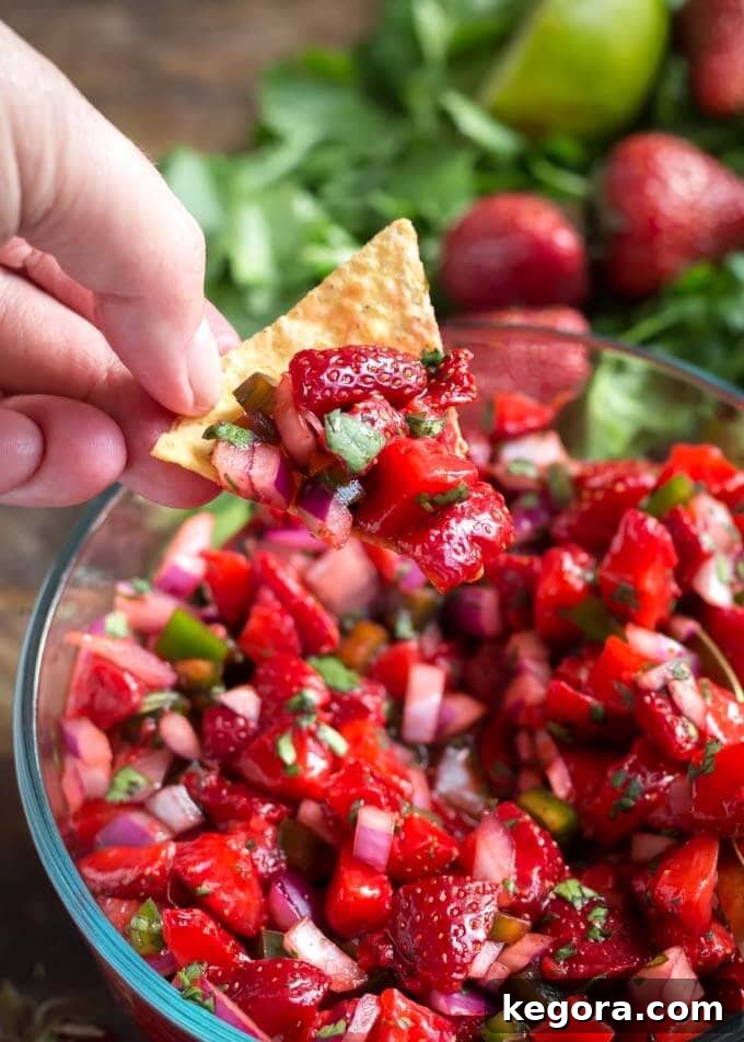 Strawberry Zing Salsa 3 Hand using tortilla chip to scoop up Fresh Strawberry Salsa in glass bowl, highlighting the enticing texture and readiness for enjoyment