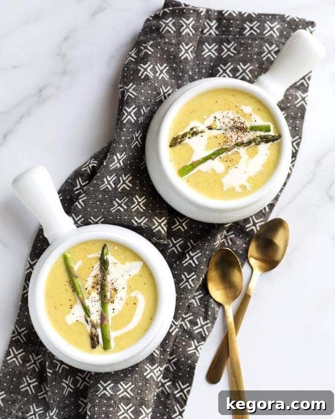 Two white bowls of creamy Instant Pot Asparagus Soup garnished with fresh black pepper, set on a black patterned napkin with two golden spoons
