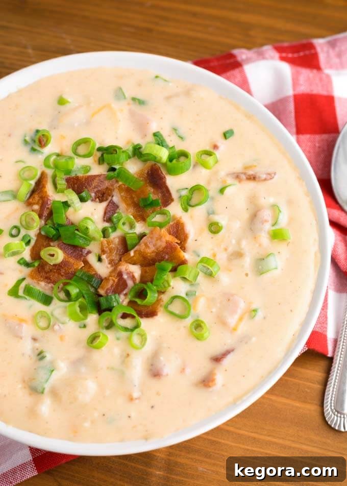 Close-up of a white bowl filled with creamy Slow Cooker Loaded Potato Soup, generously garnished with crispy bacon, shredded cheddar, and vibrant green onions, resting on a charming red gingham napkin