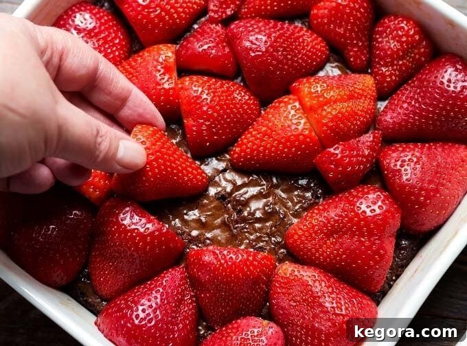 Hand placing strawberries on top of brownie in white baking dish