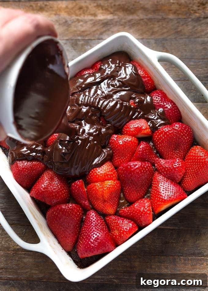 hand pouring chocolate ganache over strawberries and brownie in baking dish