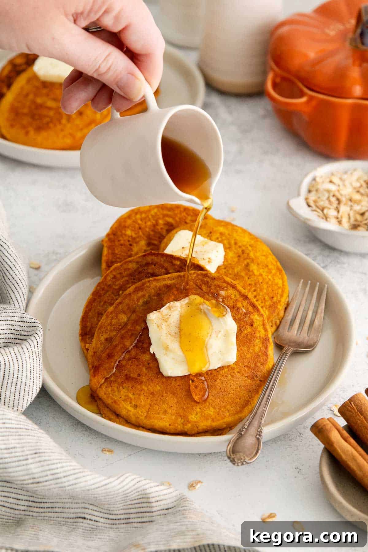 Spiced Oat Flour Pumpkin Pancakes 3 A beautiful stack of three golden oat flour pumpkin pancakes, adorned with a pat of butter and glistening with pure maple syrup being generously poured over them.