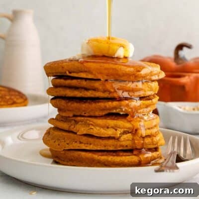 A high-angle shot of a mouthwatering stack of oat flour pumpkin pancakes, generously topped with a dollop of melting butter and a cascading pour of golden maple syrup.