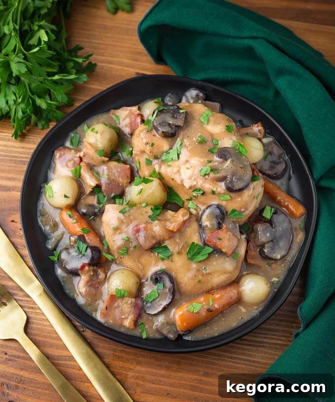 Effortless Slow Cooker Coq Au Vin 2 Coq Au Vin served in a black bowl on a rustic wooden board, accompanied by a gold fork and knife, highlighting its elegant presentation.
