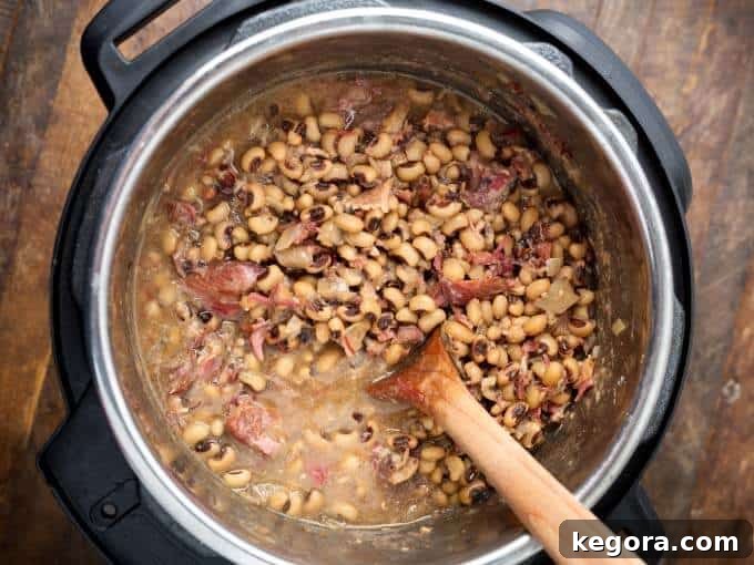 Close-up of Southern Black Eyed Peas simmering in an Instant Pot, with a wooden mixing spoon resting inside, highlighting the rich texture and steam.