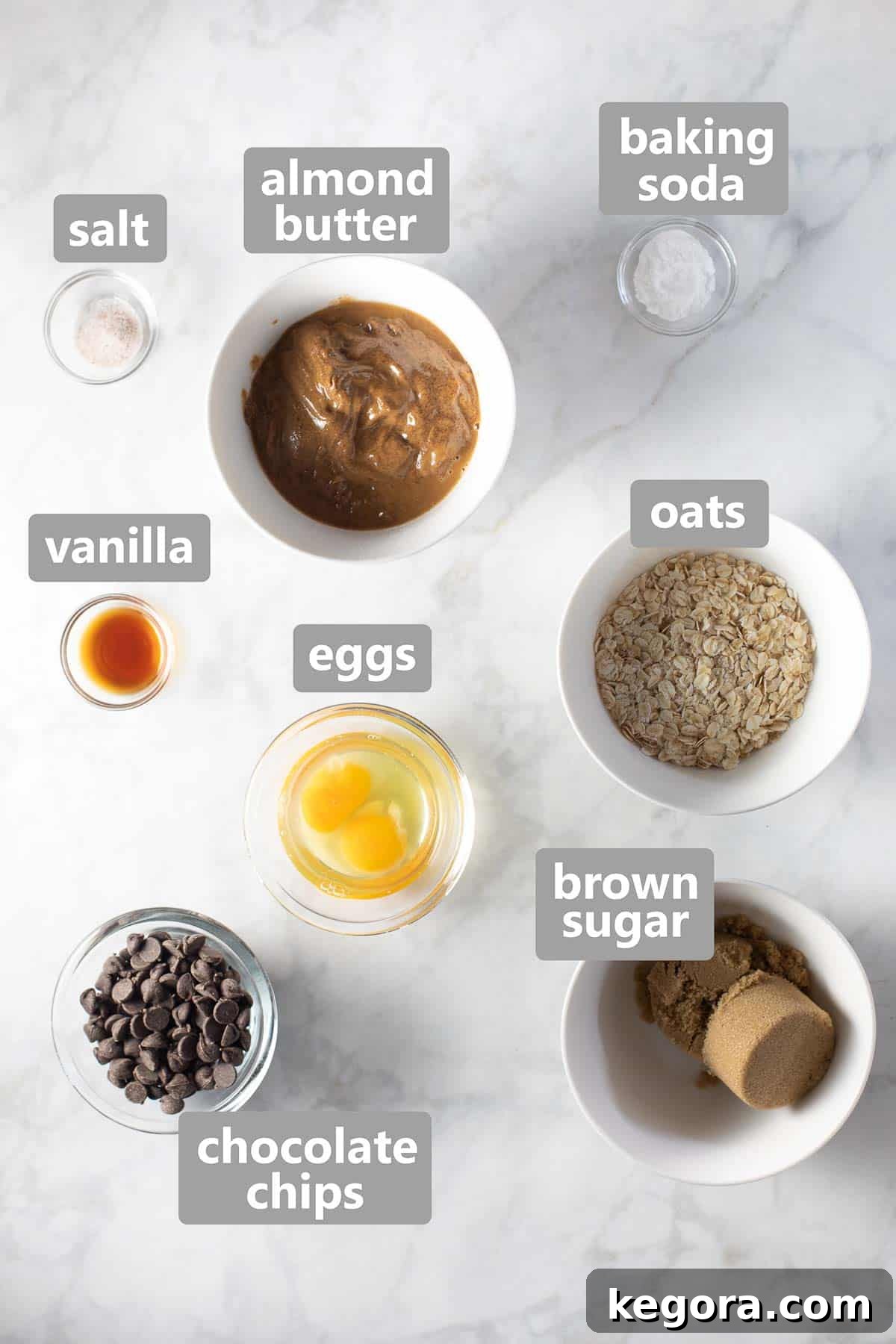 Wholesome Flourless Oatmeal Almond Butter Cookies 4 Bowls of almond butter, oats, chocolate chips, brown sugar, and eggs, neatly arranged on a marble background, ready for baking.
