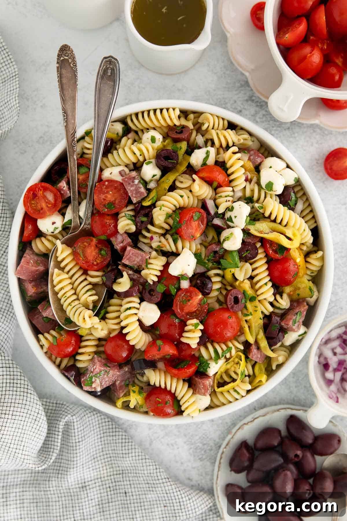 Vibrant Gluten-Free Italian Pasta Salad 3 pasta salad in a white bowl with two spoons and ingredients in the background.