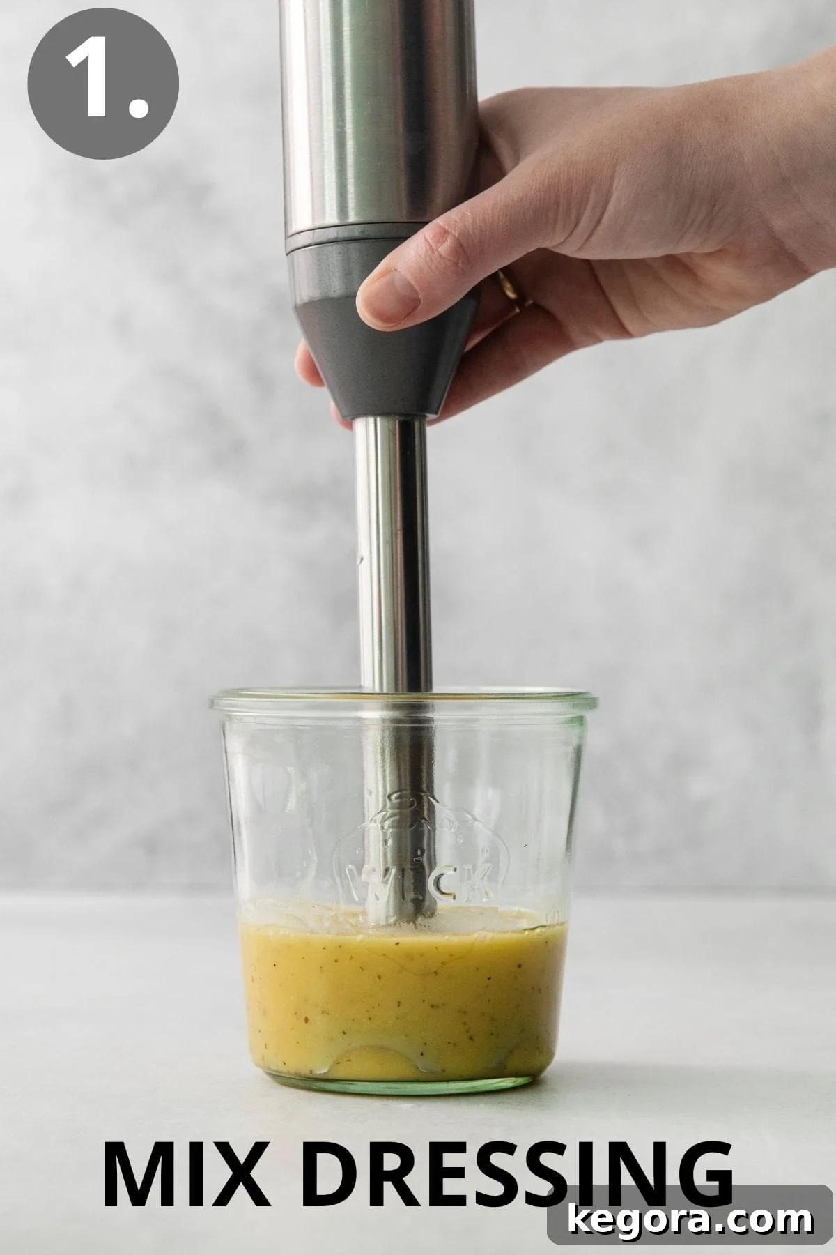 A hand using an immersion blender to mix ingredients for salad dressing in a jar