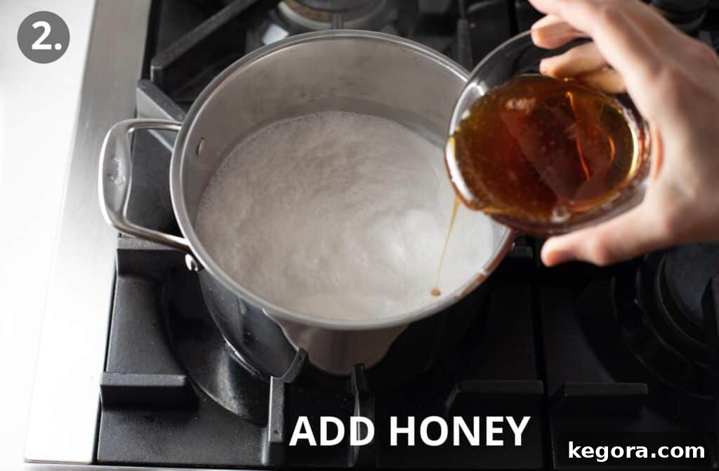 Simple Vegan Fudge 5 Liquid honey being whisked into simmering coconut milk in a metal pot on the stove, continuing the reduction process for the fudge base.