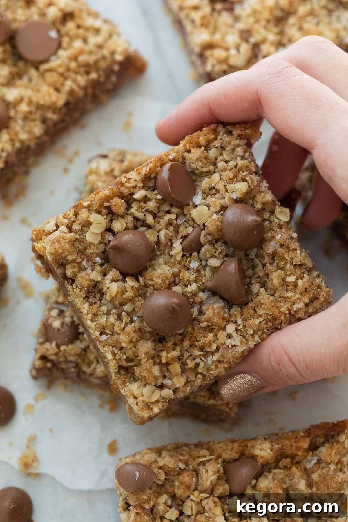 A hand reaching for a single gluten-free chocolate caramel oatmeal bar, showcasing its layers and tempting appearance.