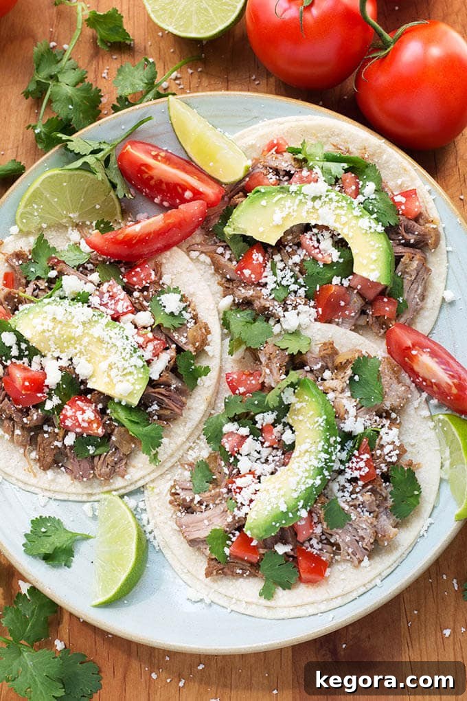Three Instant Pot Pork Carnitas tacos on corn tortillas on a white plate garnished with fresh lime wedges and chopped tomatoes.