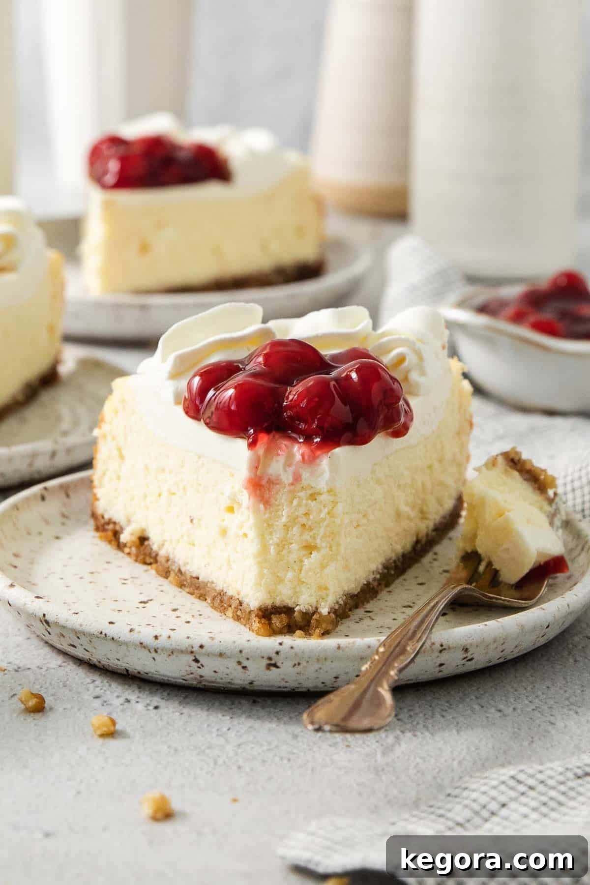 A close-up photograph of a slice of gluten-free cheesecake on a plate, adorned with whipped cream and cherries.