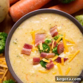 Loaded Broccoli Cheese & Potato Soup in a dark colored bowl with carrots and broccoli in the background, garnished with fresh herbs