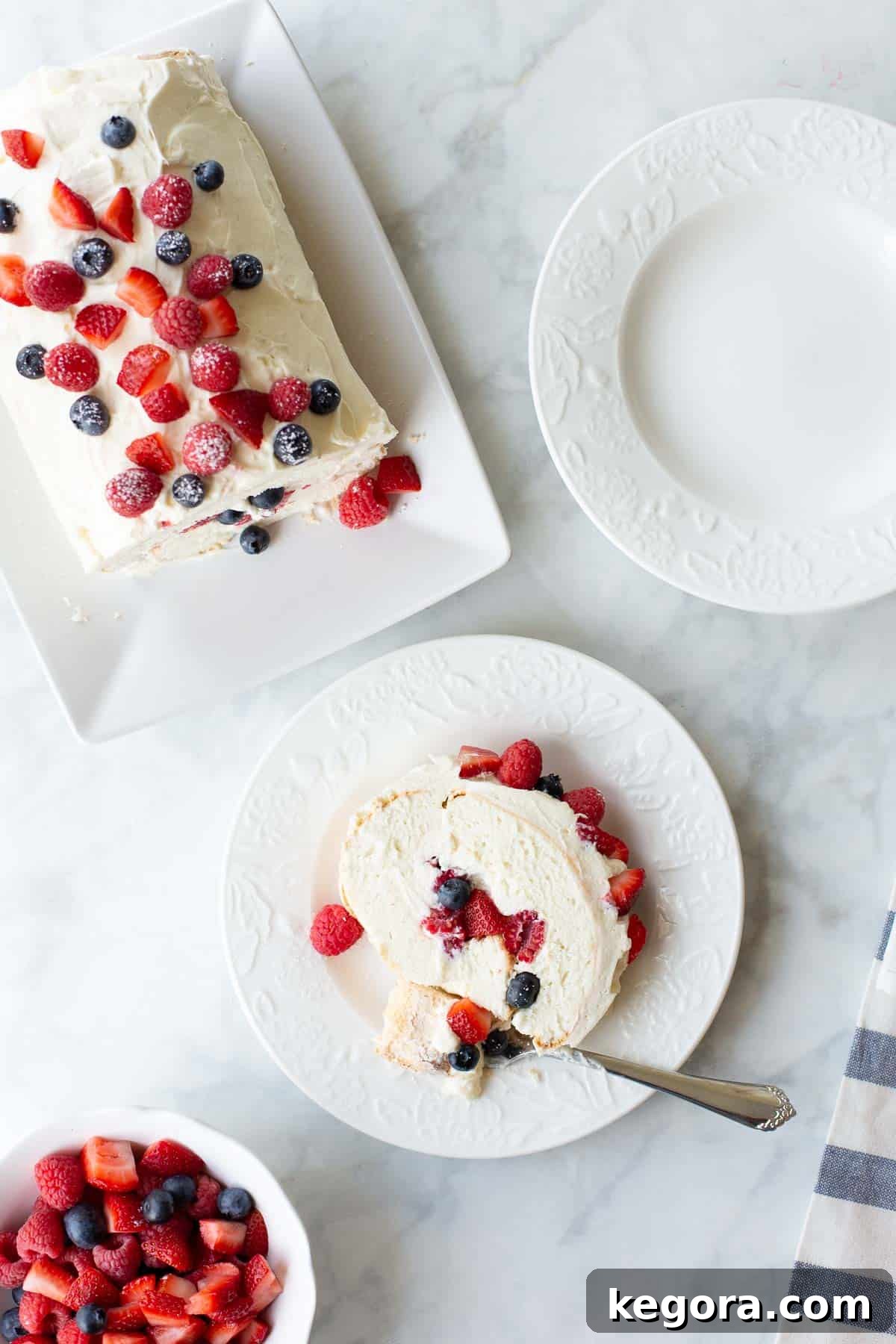 Overhead shot of a perfect slice of gluten-free angel food cake roll on a white plate, served alongside a bowl of fresh berries.
