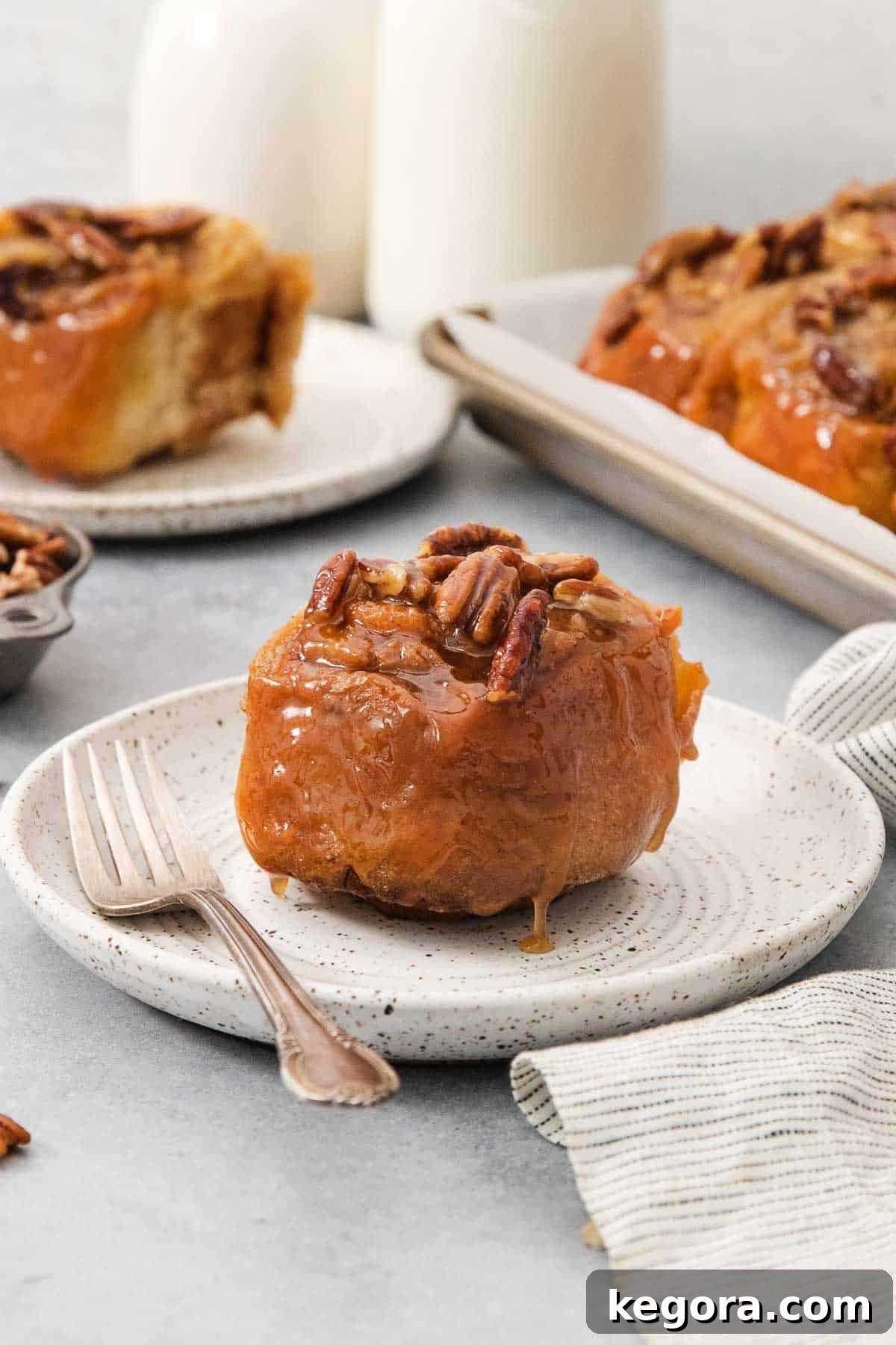 Decadent Gluten-Free Sticky Buns 3 A single gluten-free sticky bun on a white plate with a fork, with more sticky buns blurred in the background, showcasing its gooey caramel and pecan topping.
