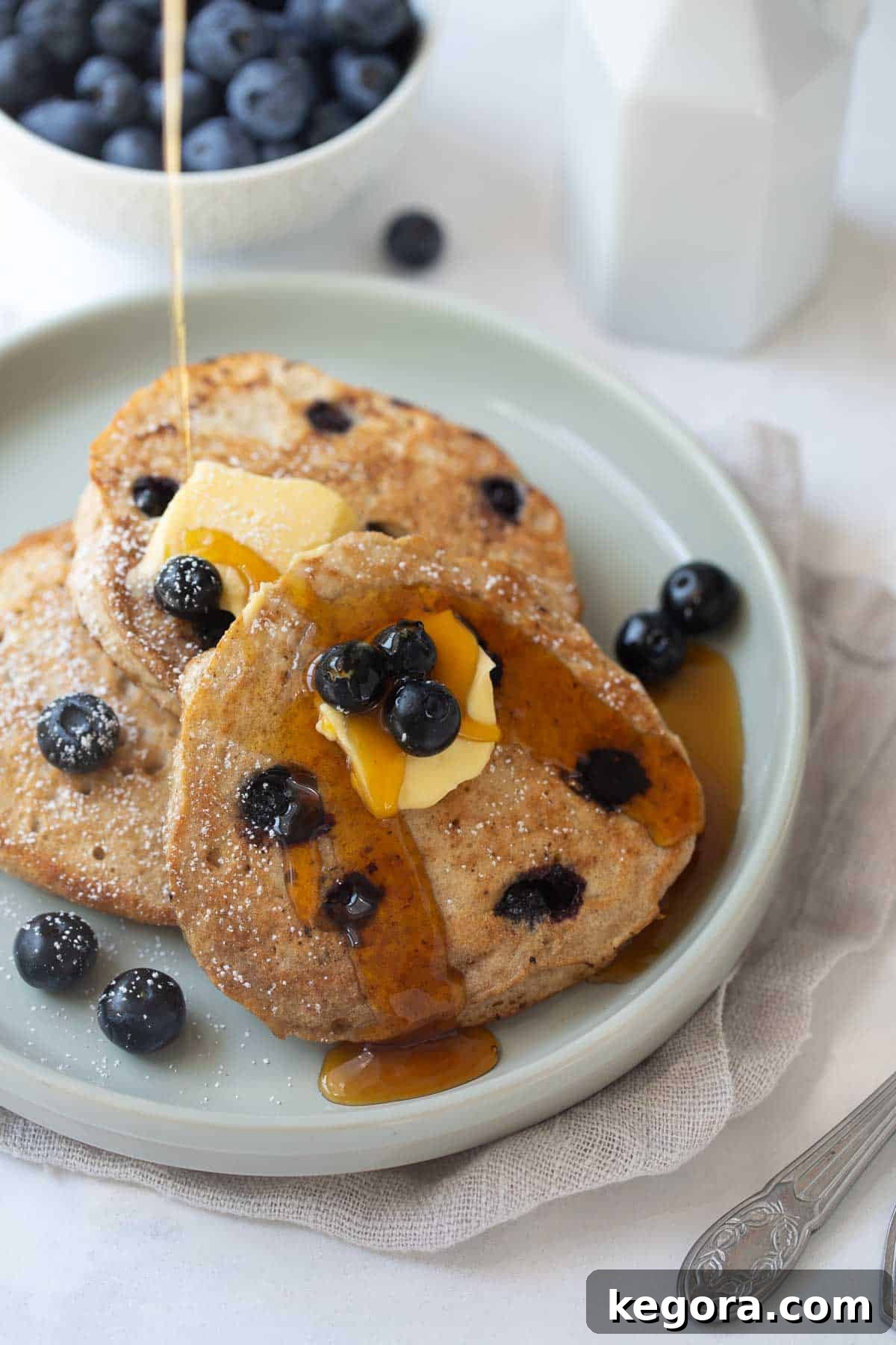 A stack of golden-brown buckwheat blueberry pancakes on a gray plate, topped with a pat of butter and drizzled with maple syrup, surrounded by fresh blueberries on a white background.