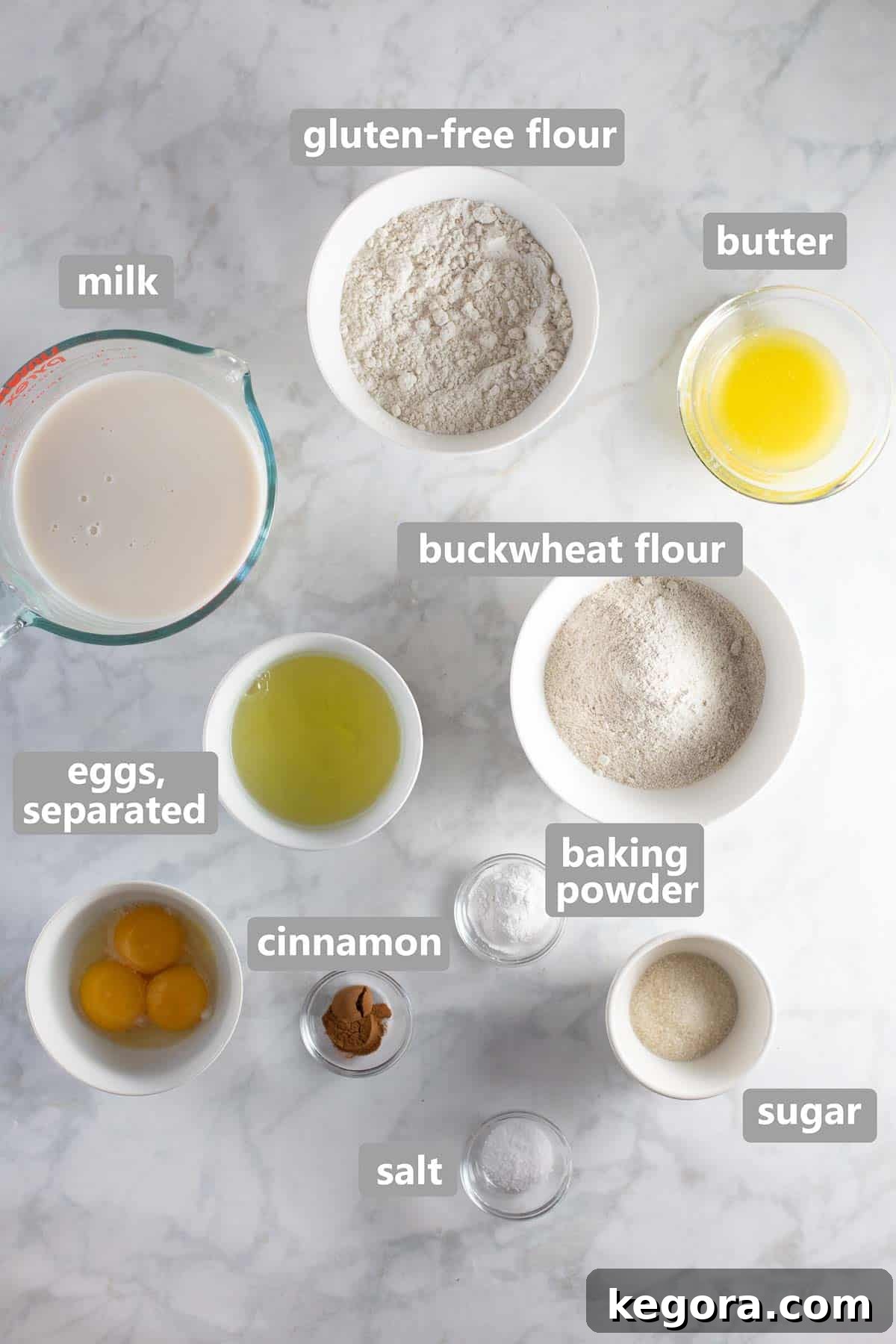 Overhead shot of all dry and wet ingredients for buckwheat pancakes laid out in individual bowls on a white table