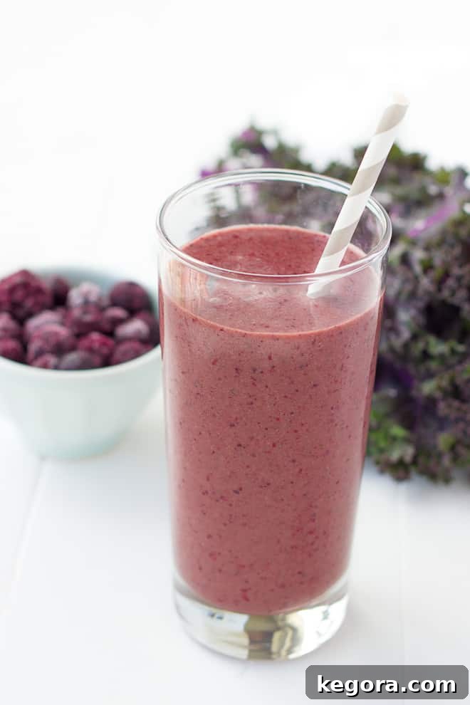 A beautiful side view of a purple green smoothie in a glass, with a bowl of berries softly blurred in the background.