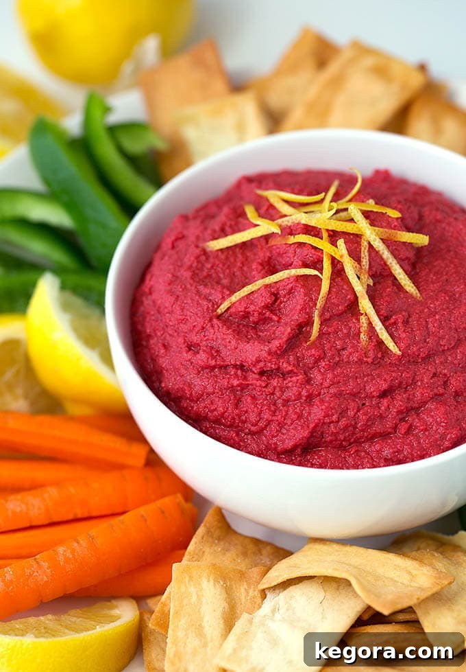 Crimson Beet Hummus 2 A serving of Red Beet Hummus in a white bowl, garnished with olive oil and surrounded by an assortment of colorful dippers like pita chips, carrot sticks, green bell pepper sticks, and lemon wedges.