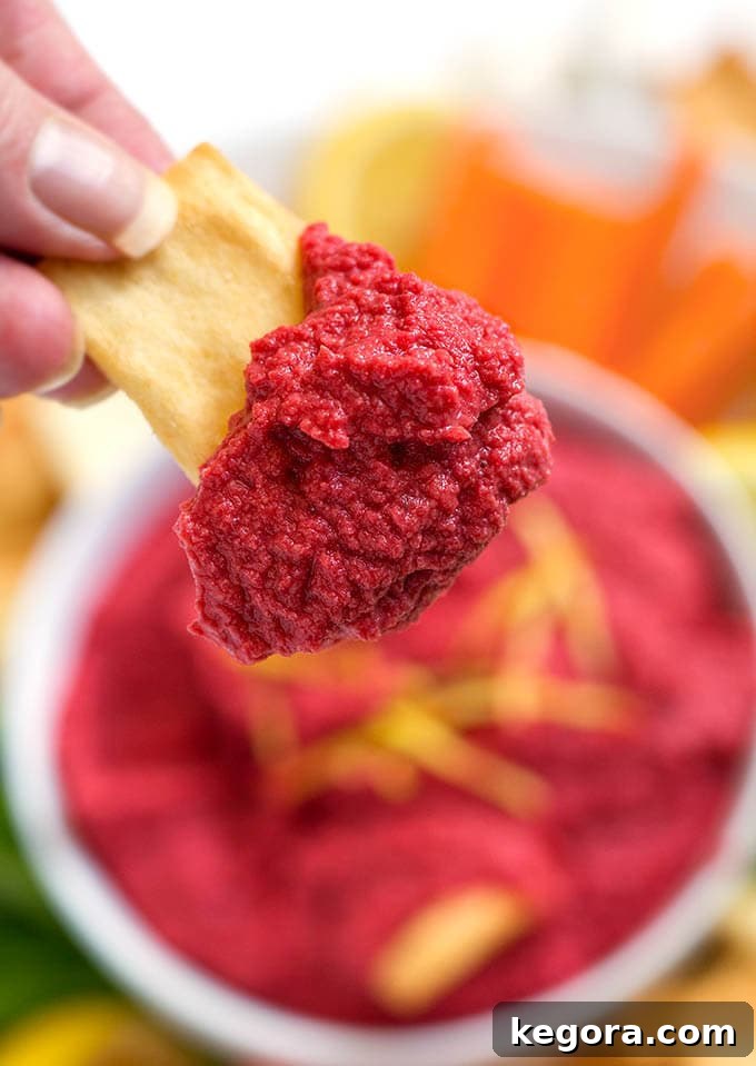 Crimson Beet Hummus 3 Close-up shot of a human hand holding a pita chip with a generous dollop of creamy, vibrant Red Beet Hummus.