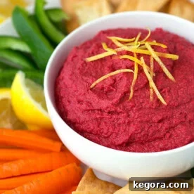 Crimson Beet Hummus 4 Red Beet Hummus in a white bowl surrounded by pita chips carrot sticks green pepper sticks and lemon wedges