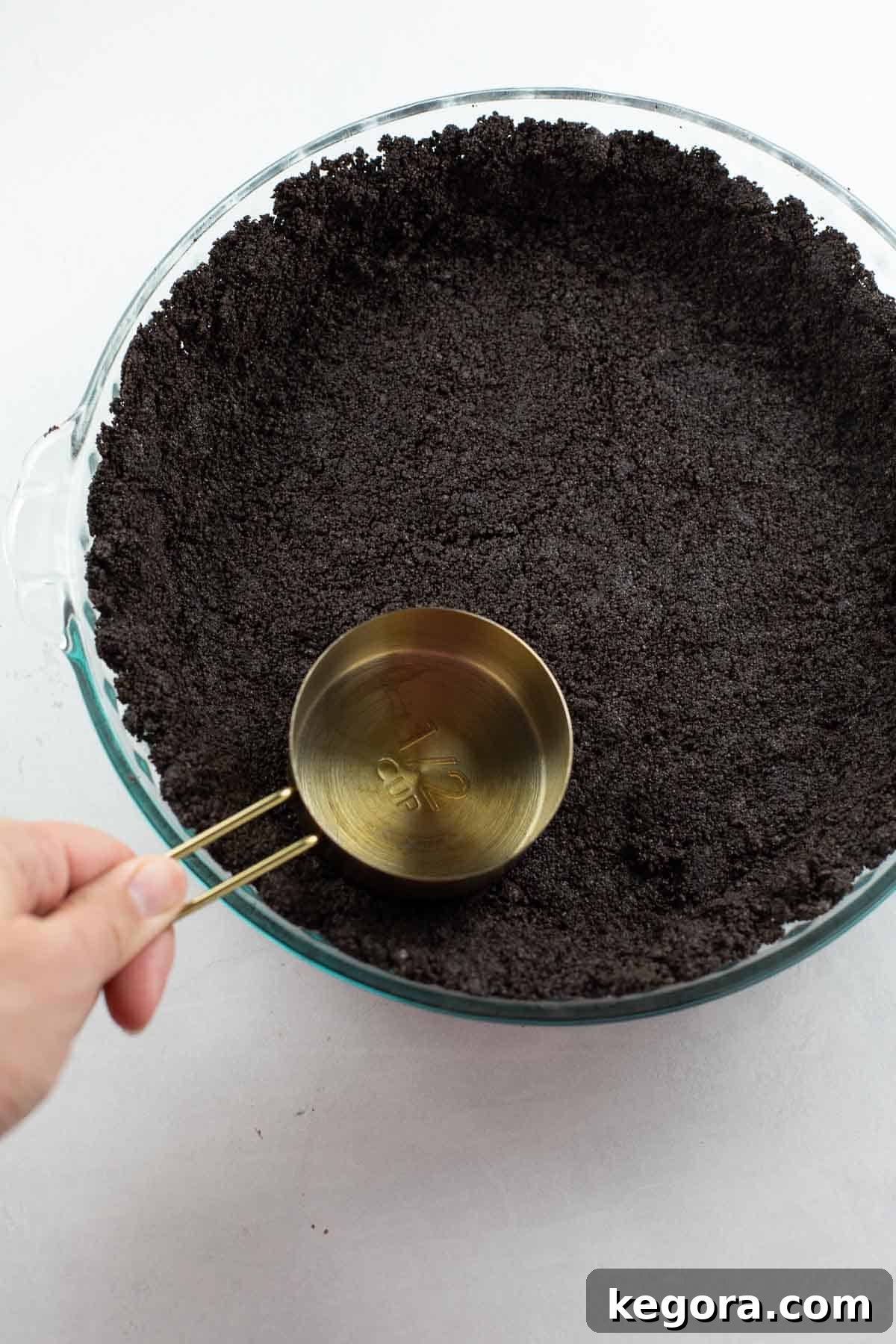 A hand using a measuring cup to press gluten-free Oreo pie crust into a pie plate, ensuring an even base.
