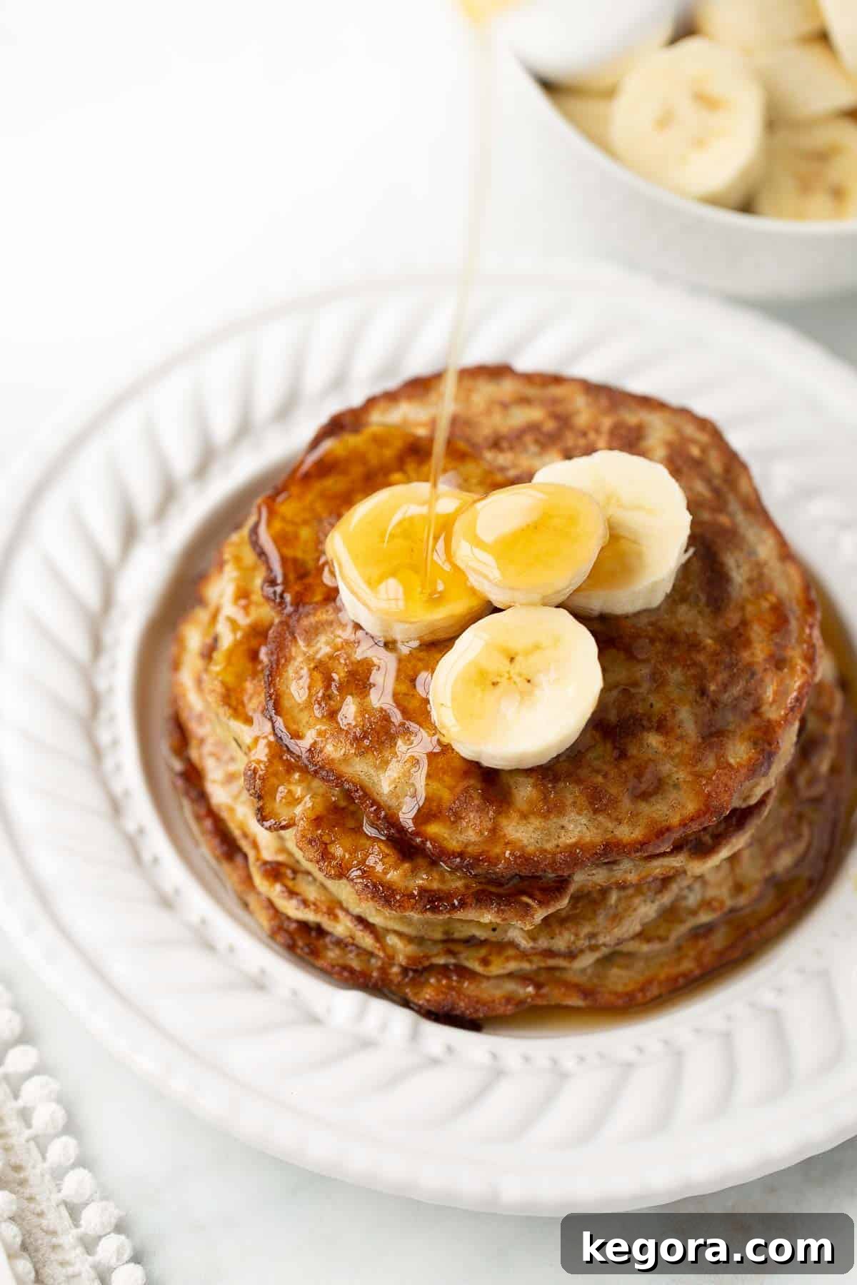 Syrup being poured generously over a stack of fluffy banana oat pancakes, ready to be enjoyed.
