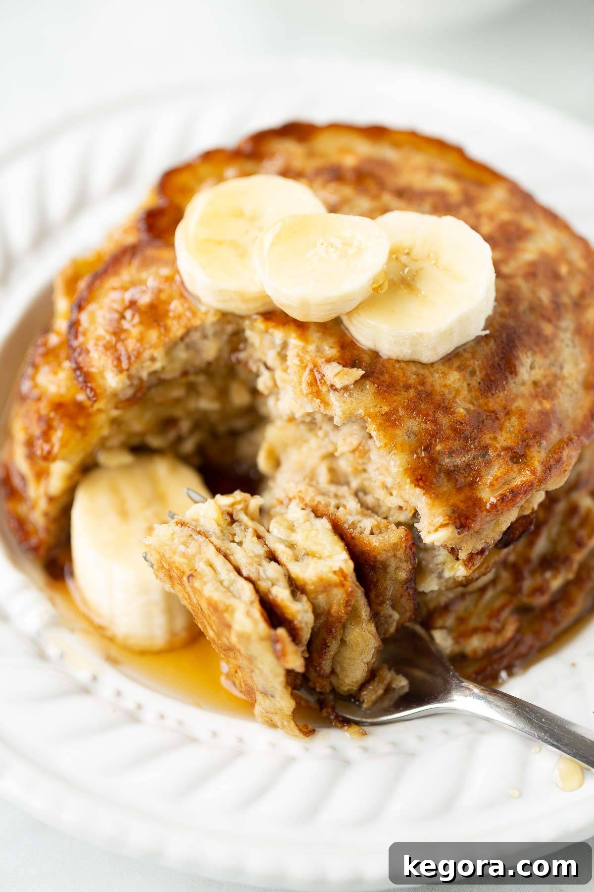 Close-up shot of a fork taking a bite of banana oat pancakes on a pristine white plate, garnished with fresh berries and banana slices.