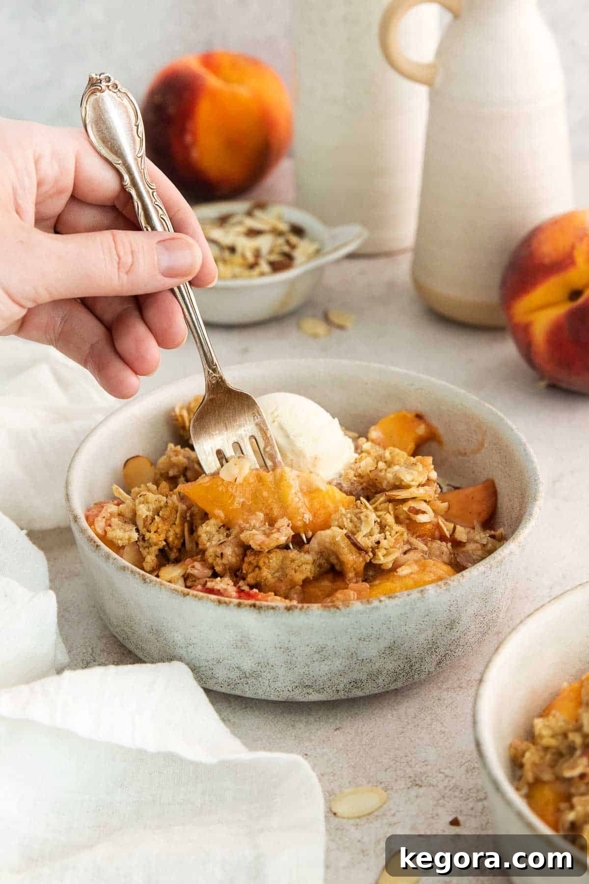 A close-up shot of a bowl of peach crisp with vanilla ice cream, with a hand holding a fork, ready to take a bite.