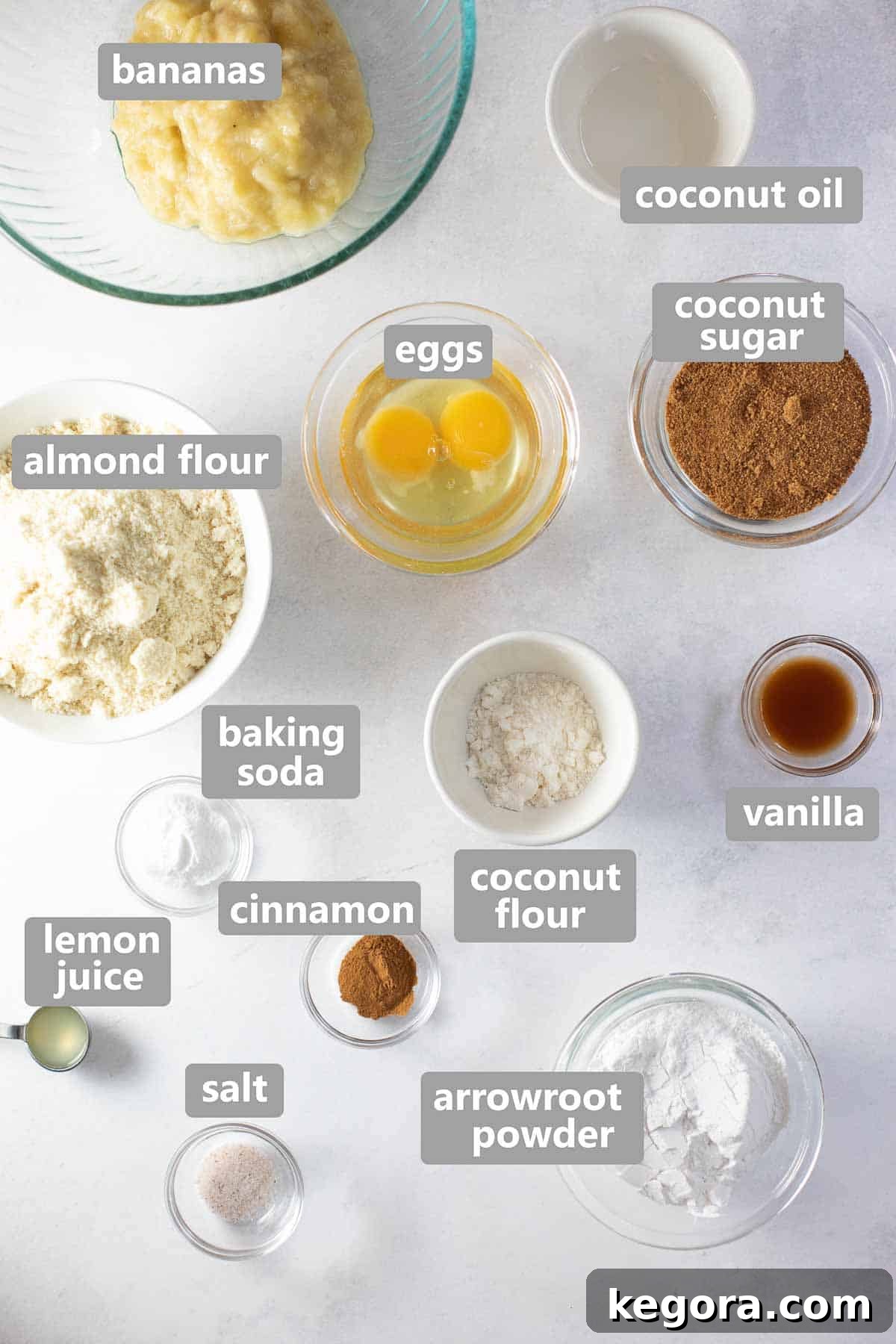 Overhead shot of ingredients to make paleo banana bread, including mashed bananas, eggs, flours, and spices, set out in bowls on a wooden surface.