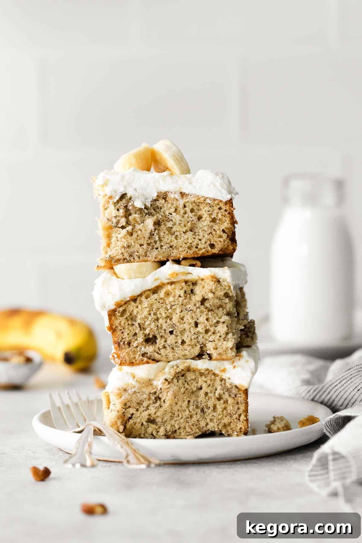 Golden Gluten-Free Banana Treat 3 Moist gluten-free banana cake slices stacked on top of each other on a white plate, showcasing the creamy frosting.