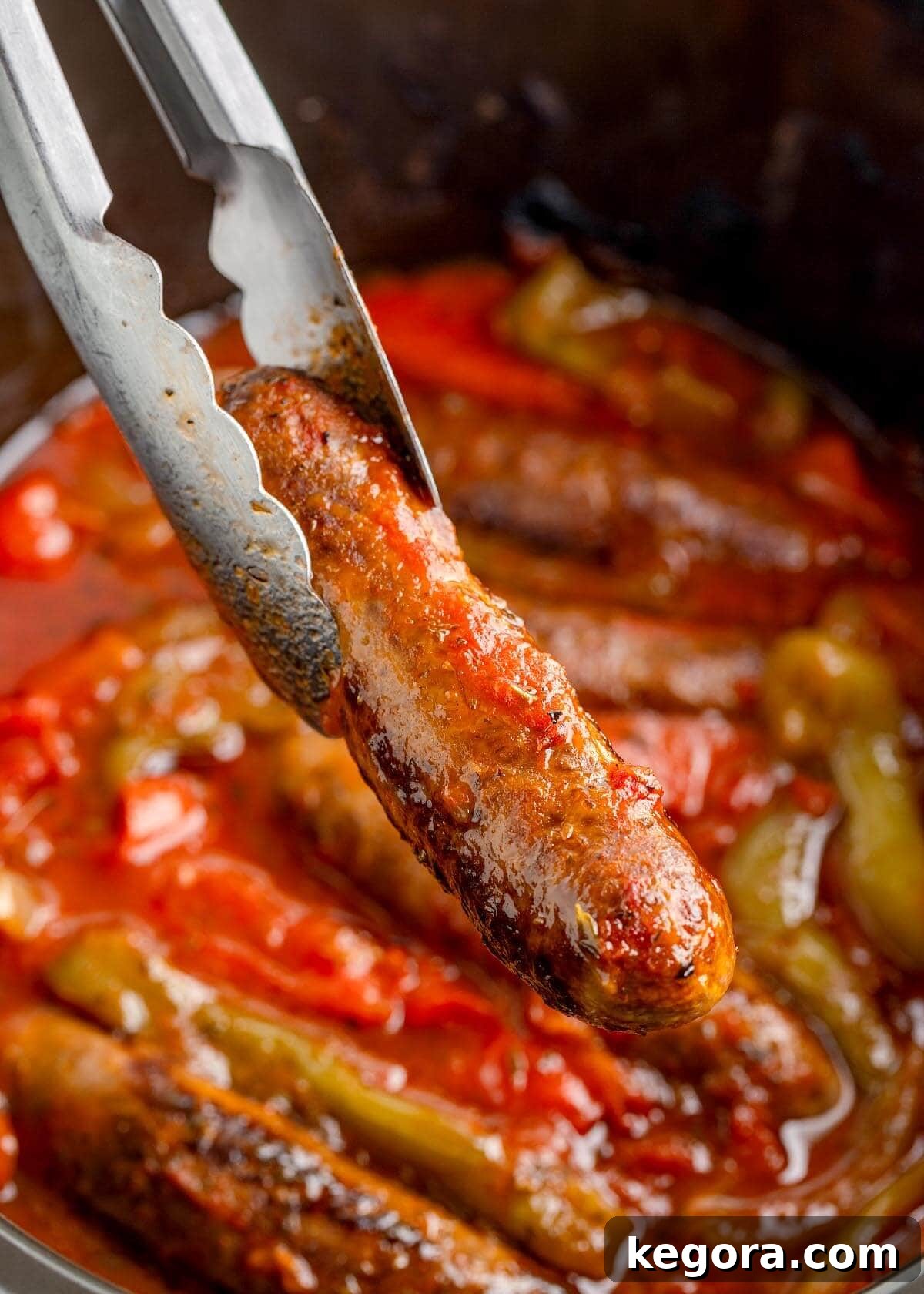 Set It and Forget It Sausage and Peppers 8 Tongs holding a perfectly cooked Italian sausage link over the slow cooker filled with vibrant peppers and onions.