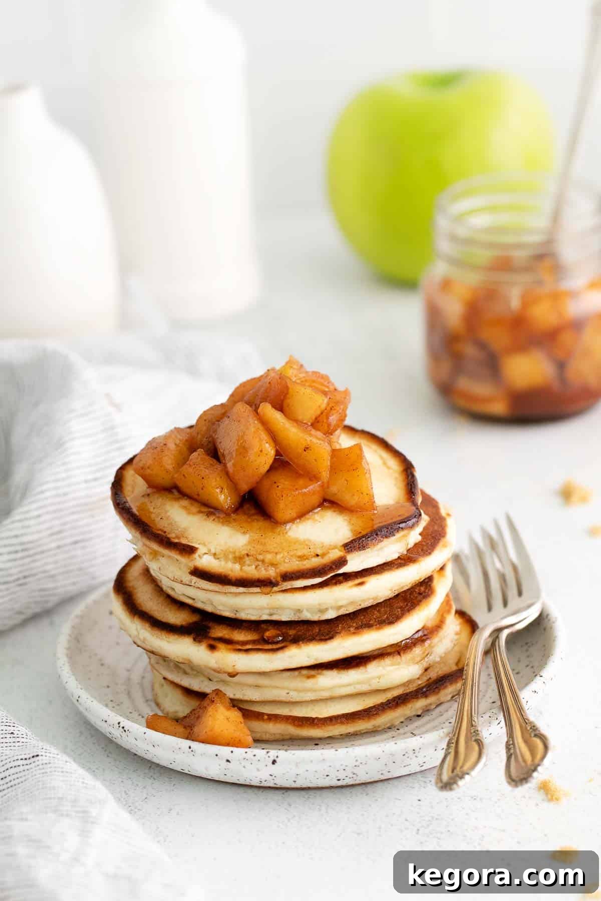 Rustic Apple Compote 6 Fluffy pancakes topped with generous spoonfuls of apple compote, with a fork nearby and a jar of compote in the background.