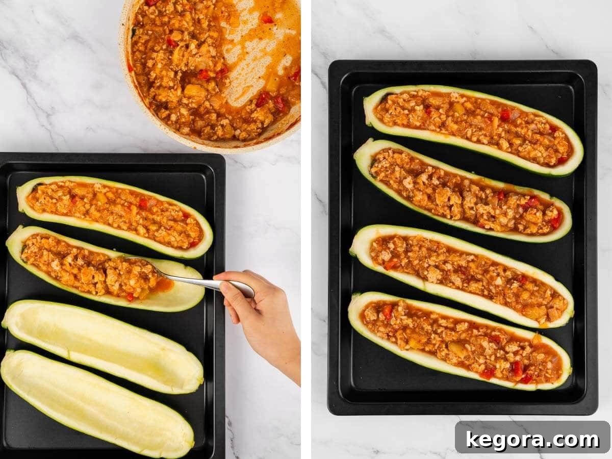A hand using a spoon to generously stuff the zucchini boats with the savory chicken and vegetable filling, with four stuffed zucchini boats ready.