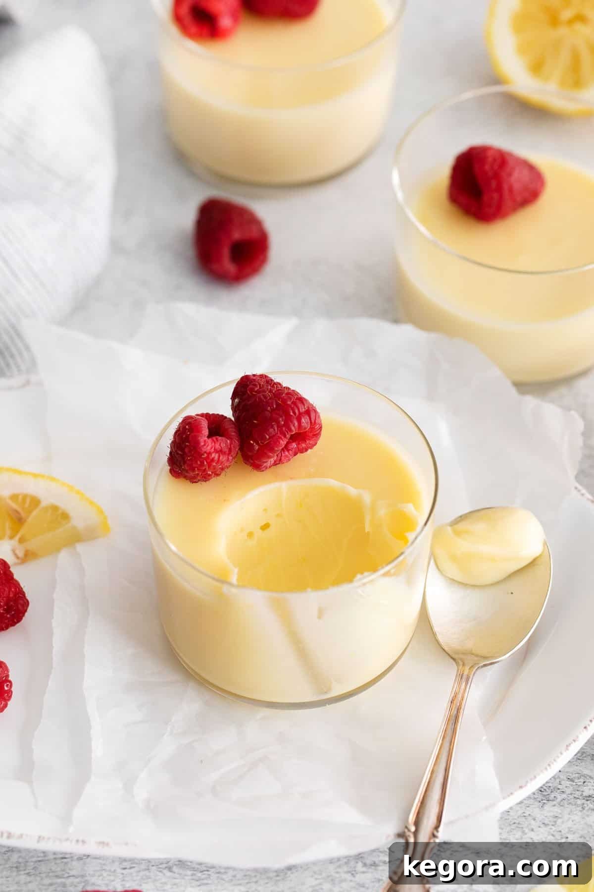 A single serving of lemon posset in a glass dish, garnished with raspberries, with a spoon having taken a bite, highlighting its creamy texture.