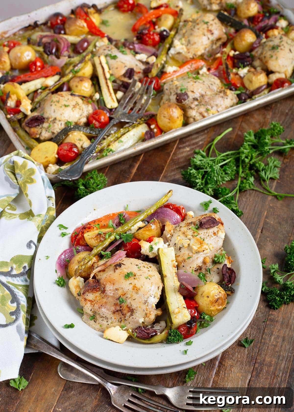 One-Pan Greek Lemon Herb Chicken 2 Greek Chicken Dinner on a plate, sheet pan of food behind.