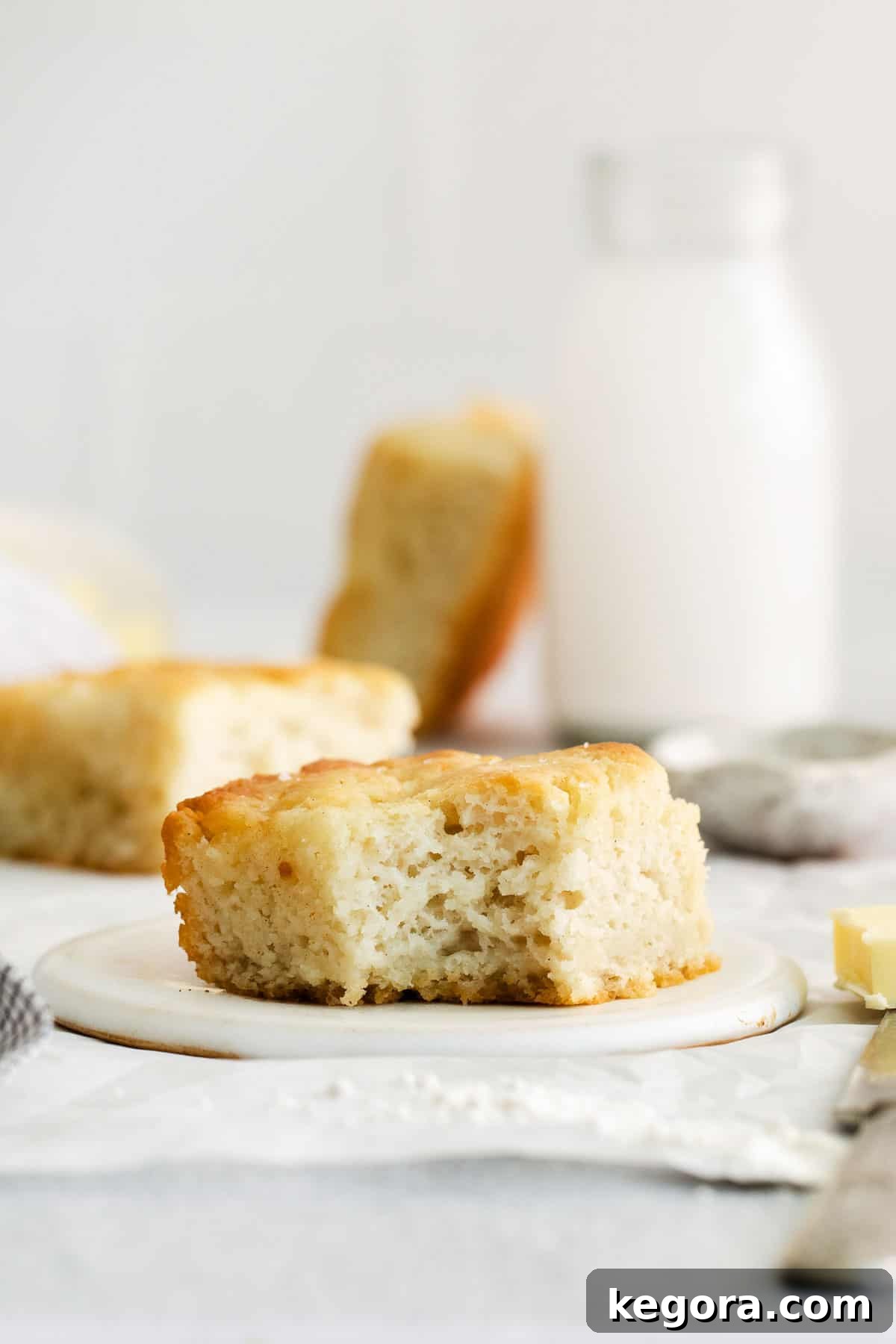 Butter-Dipped Gluten-Free Biscuits 3 A single gluten-free biscuit on a white plate, with a glass of milk and a baking dish of more biscuits blurred in the background.
