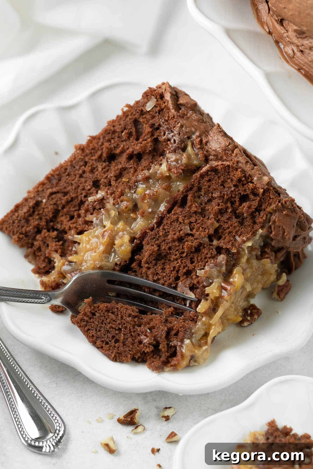Decadent Gluten-Free German Chocolate Cake 10 A close-up of a slice of Gluten-Free German Chocolate Cake on a white plate, with a fork poised to take a bite, highlighting the moist cake and rich filling.