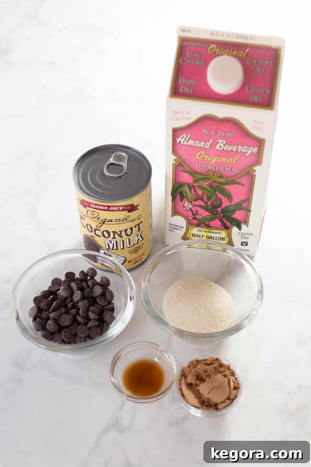 Plant-Based Cocoa Bliss 3 A flat lay of ingredients for dairy-free hot chocolate, including cartons of almond and coconut milk, a bowl of chocolate chips, cocoa powder, sugar, and vanilla extract.