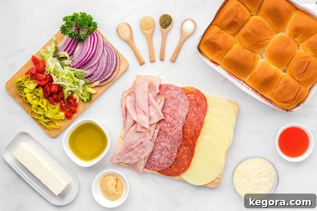 Italian Sub Sliders ingredients laid out: olive oil, vinegar, Italian seasoning, mustard, butter, parsley, garlic powder, onion powder, salt, parmesan, Hawaiian rolls, ham, salami, pepperoni, provolone, red onion, cherry tomatoes, pepperoncini, lettuce.
